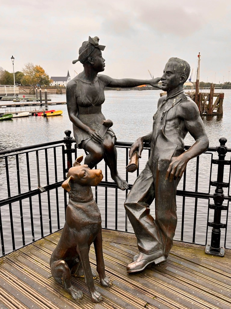 Bronze statue of a woman sitting on a railing by the water, touching the face of a standing man; a dog sits beside them — located at Cardiff Bay.