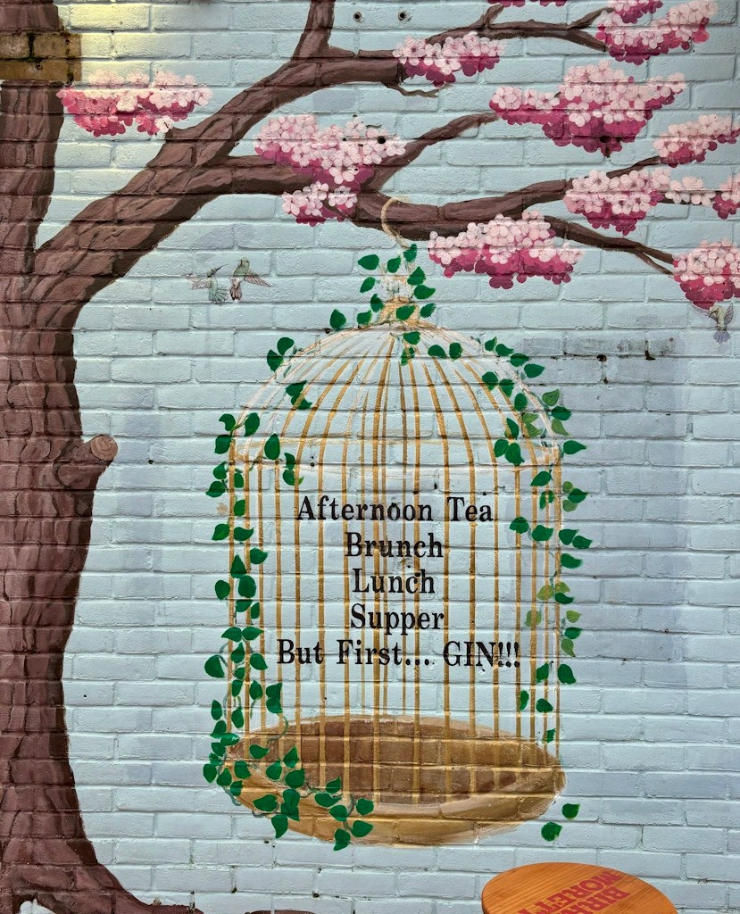 Wall mural of a golden birdcage wrapped in green vines, set against a pale blue brick wall with pink cherry blossoms; inside the cage, the text reads: ‘Afternoon Tea, Brunch, Lunch, Supper, But First… GIN!!!’.
