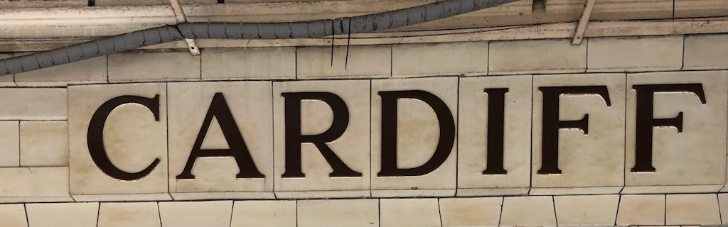 Tiled wall with the word ‘Cardiff’ in large dark letters, part of an old station sign.