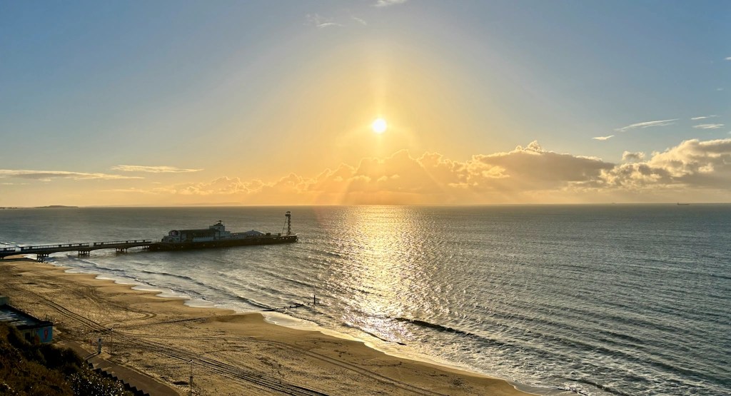 Golden sunrise over a calm sea with gentle waves, a sandy beach in the foreground, and the long Bournemouth pier extending into the water on the left side.