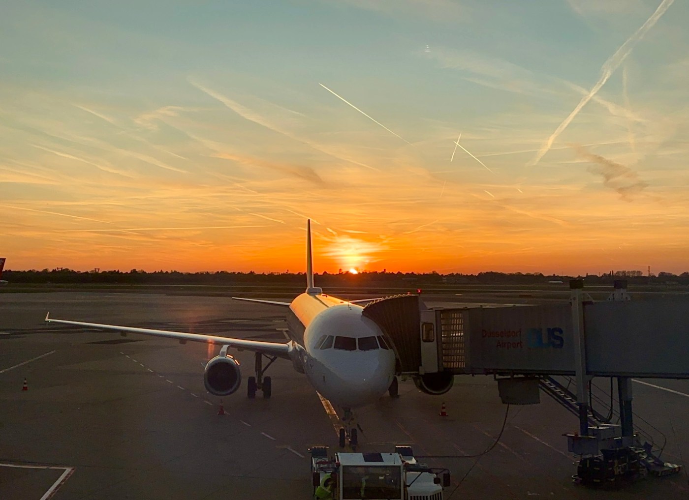 A passenger aircraft parked at the gate, connected to a jet bridge at Düsseldorf Airport, with the sun setting on the horizon and a sky filled with warm colors and crossing contrails.