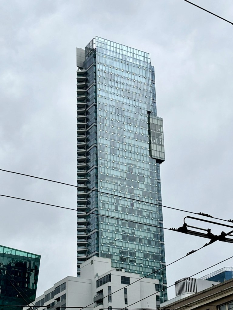Tall modern glass high‑rise building in Vancouver viewed from street level, with power lines crossing the frame and an overcast sky above.
