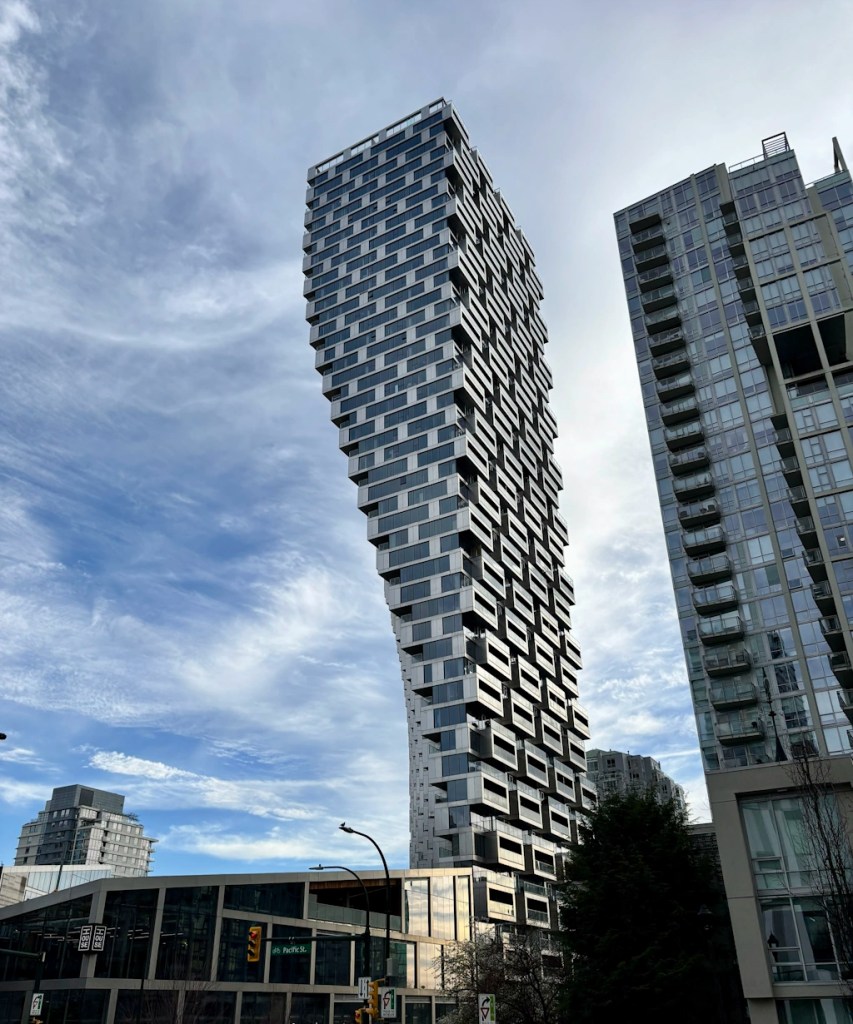 Distinctive twisting high‑rise tower with irregular stacked balconies, rising above surrounding buildings against a partly cloudy sky.