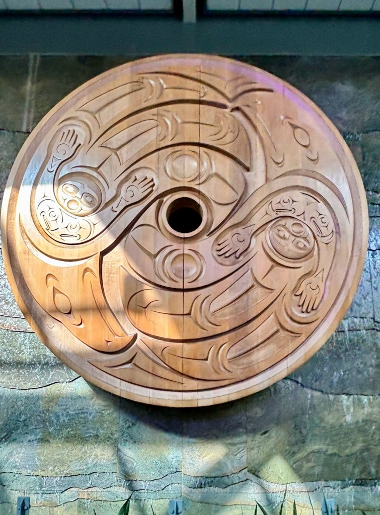 Circular carved wooden artwork with stylized Indigenous figures and flowing patterns, mounted indoors and illuminated from above.