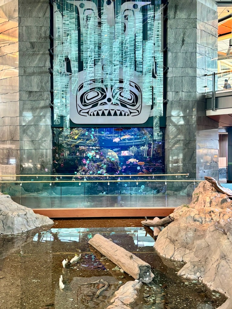 Indoor water feature at Vancouver International Airport with rocks and driftwood in the foreground, and a glass wall revealing an aquarium and Indigenous design motif.