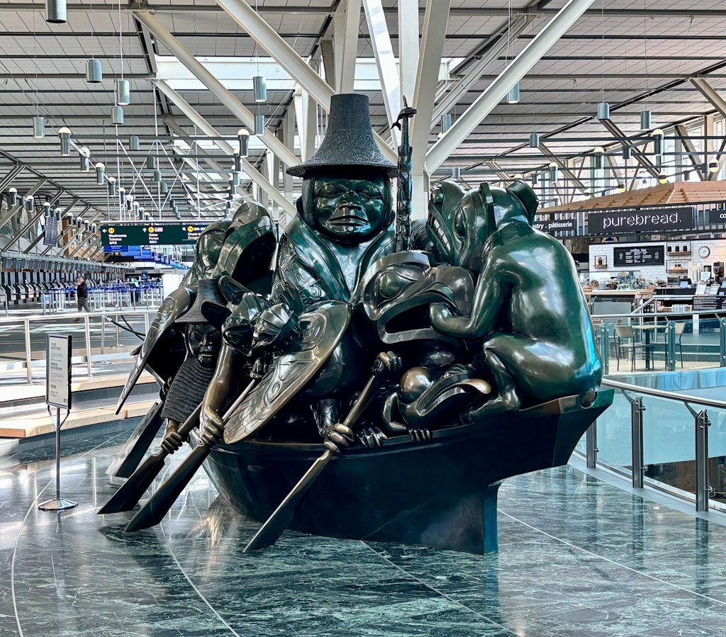 Dark bronze Haida sculpture displayed in Vancouver International Airport, depicting multiple human and animal figures in a canoe within a spacious terminal hall.