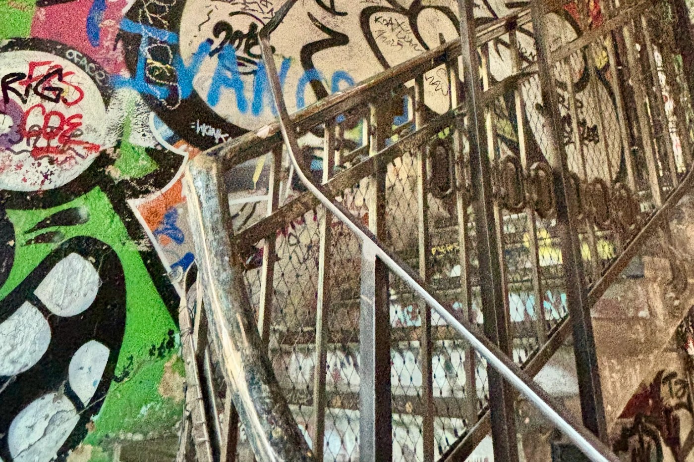 Close-up view of a metal staircase behind a wire-mesh fence, covered in colorful graffiti. The concrete wall features layered street art in green, blue, red, and black, while the metal railings and steps create strong diagonal lines across the image, giving an urban, gritty atmosphere.