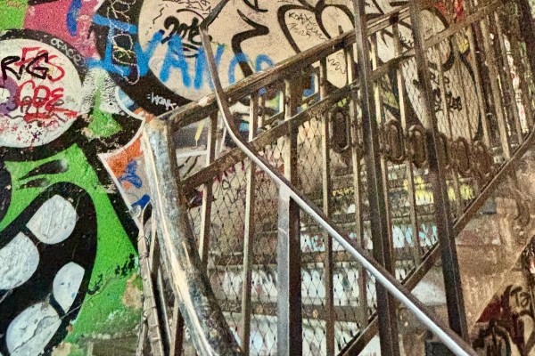 Close-up view of a metal staircase behind a wire-mesh fence, covered in colorful graffiti. The concrete wall features layered street art in green, blue, red, and black, while the metal railings and steps create strong diagonal lines across the image, giving an urban, gritty atmosphere.