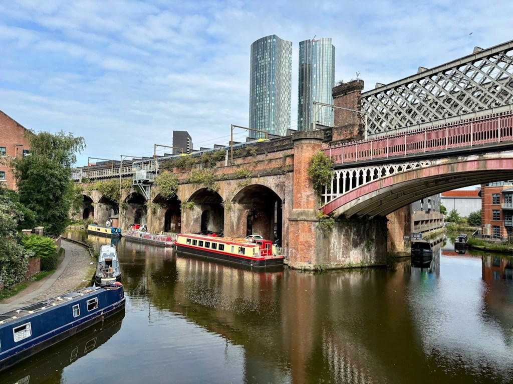 Manchester canal scene featuring a historic brick railway viaduct and an ornate iron bridge crossing the water. Several narrowboats are moored along the canal beneath the arches, while modern glass skyscrapers rise in the background. Reflections of the bridge and boats appear on the calm water, illustrating the blend of Manchester's industrial heritage and contemporary skyline.