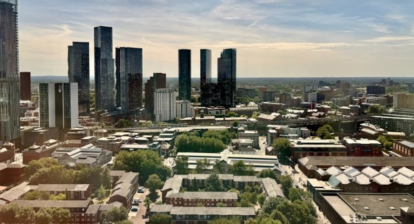 Panoramic view of Manchester's skyline from an elevated vantage point. Several tall glass skyscrapers dominate the city centre, surrounded by lower residential, commercial, and industrial buildings. Trees and green spaces are scattered throughout the urban landscape, while soft afternoon light and a partly cloudy sky stretch across the horizon.