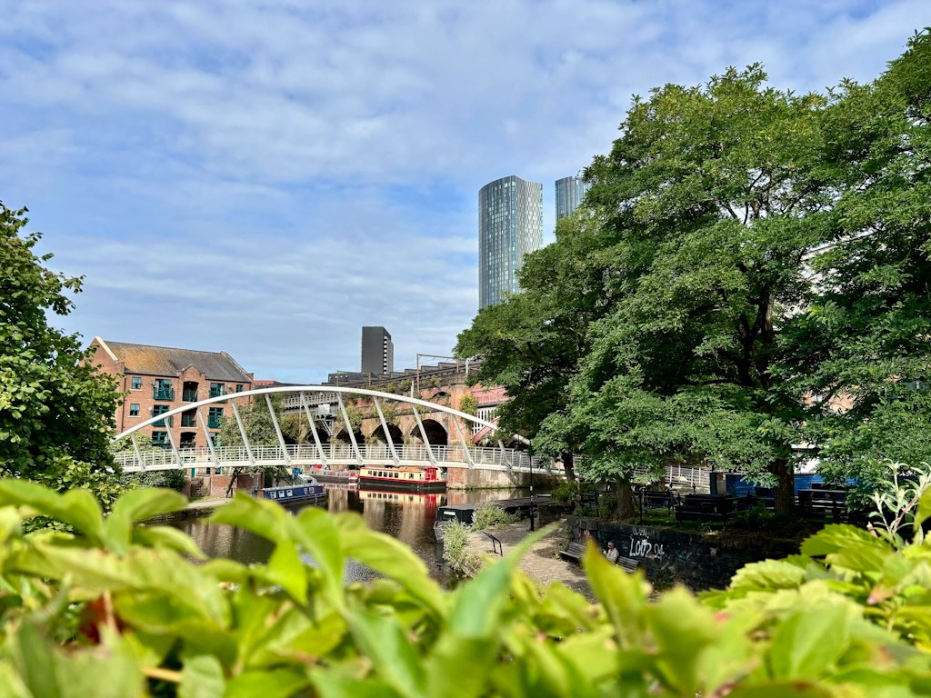 A white steel pedestrian bridge spans a canal in Manchester, framed by green foliage in the foreground. Narrowboats are moored along the waterway, while a historic brick railway viaduct and modern glass skyscrapers rise in the background. Mature trees line the canal, and a partly cloudy sky completes the scene, highlighting the contrast between Manchester’s industrial heritage and contemporary architecture.