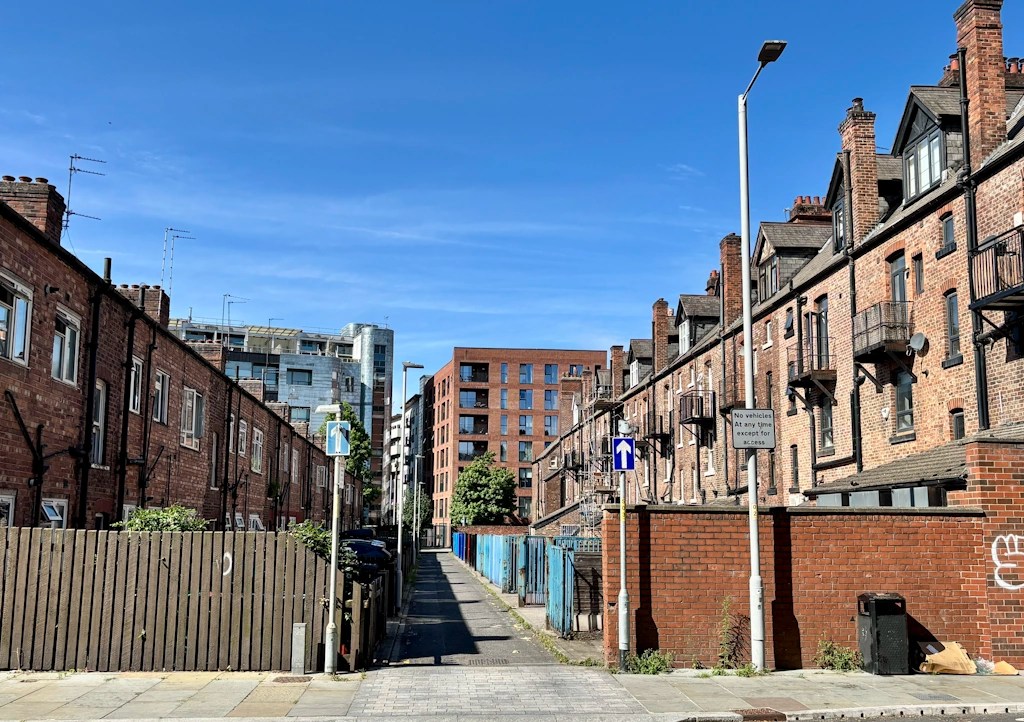 A narrow side street in Manchester lined with rows of red-brick residential buildings and modern apartment blocks in the distance. Small balconies, rear walls, utility poles, and street signs border the roadway, while the clear blue sky and bright sunlight create strong shadows along the pavement. The scene reflects the mix of historic housing and ongoing urban redevelopment found throughout the city.